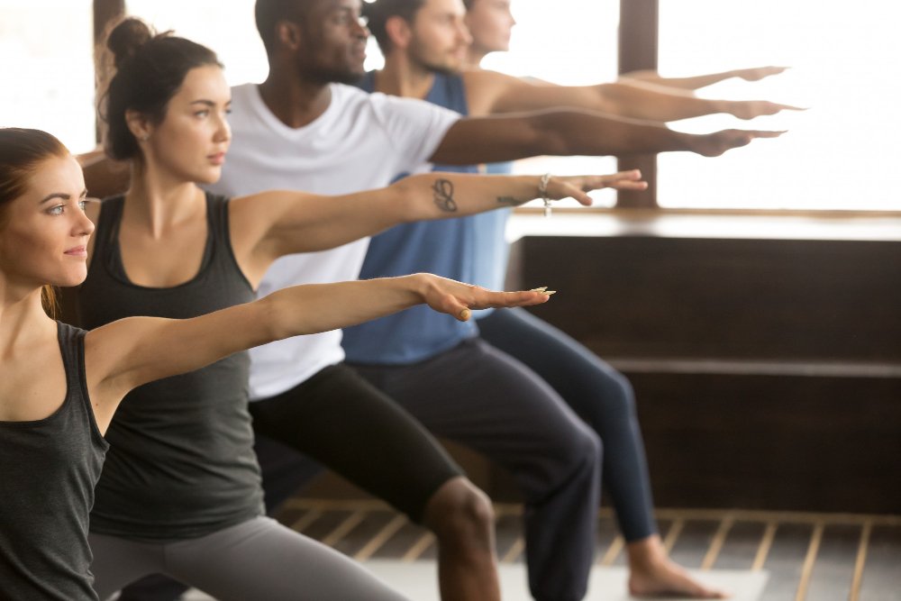 A group of people in athletic wear practice yoga together indoors, standing in a row and performing the Warrior II pose with arms extended forward and back.